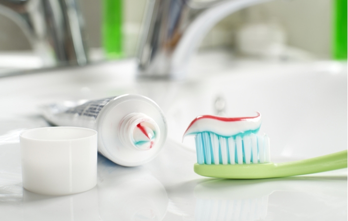 A tube of toothpaste with the cap laying next to it, beside a green toothbrush that has just been covered in the toothpaste ready for brushing, all on a bathroom counter