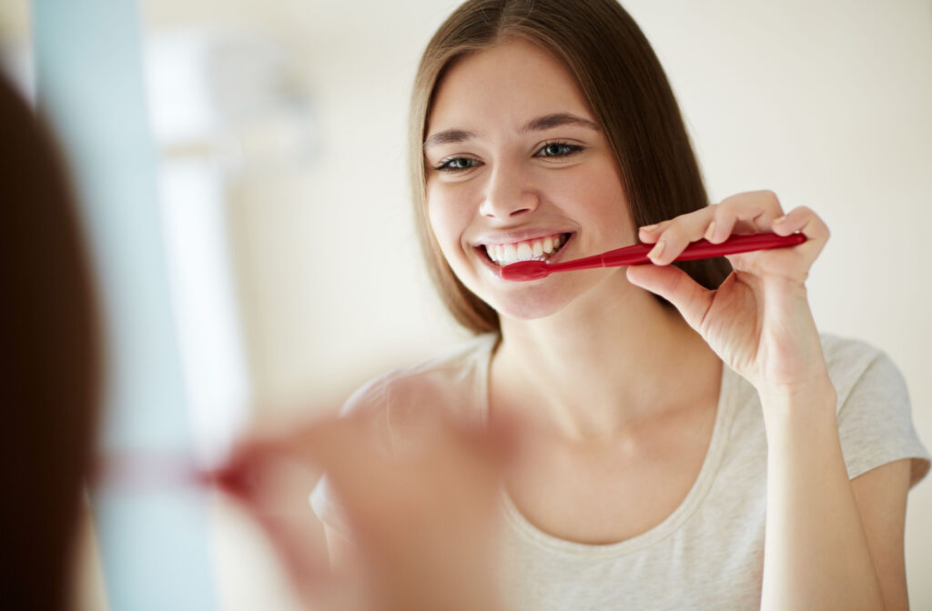 A young woman looking in the mirror and smiling while brushing her teeth with a red toothbrush.