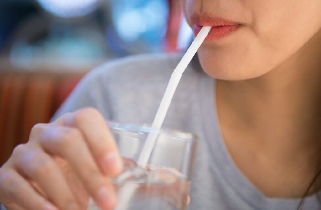 A youthful woman taking small sips of water through a straw to maintain hydration following a dental procedure.