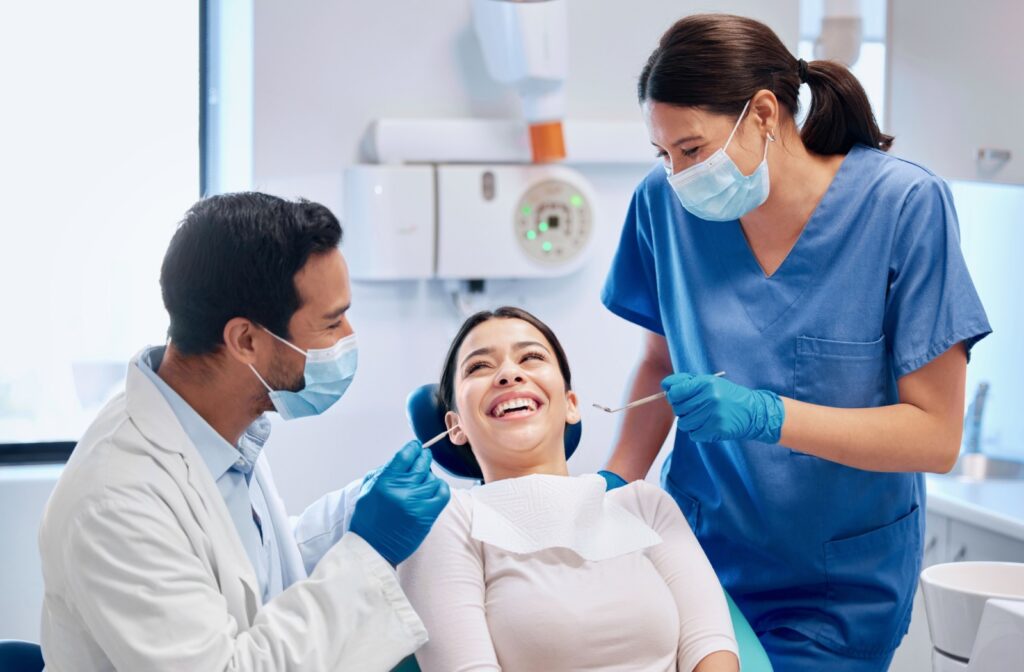 A patient lying in a dental chair smiling while a dentist and hygienist laugh with them during a dental cleaning.