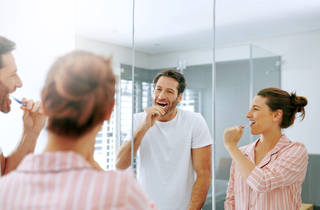 2 people brushing their teeth in the mirror to maintain good oral health, an important part of maintaining a dental implant